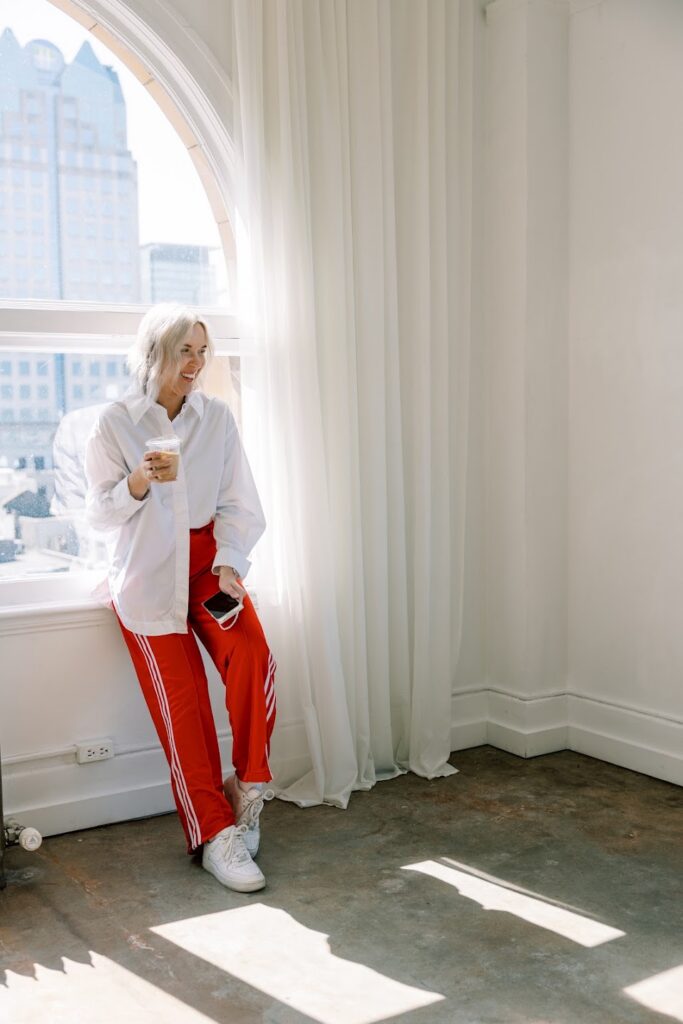 Personal Branding Coach Katrina Owens educates clients on her 90-Day Visibility Sprint Strategy stands in a white room in a white shirt and red pants drinking a coffee