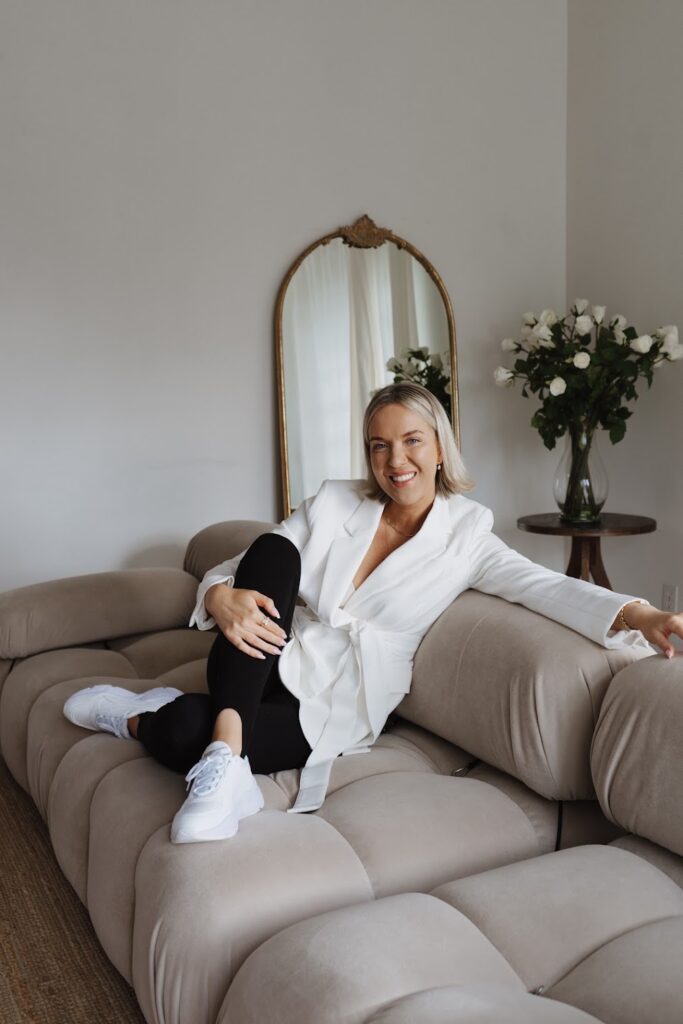 Katrina Owens, who teaches all about landing personal branding partnerships, sits on a nude couch wearing white sneakers, black pants, and a white blazer
