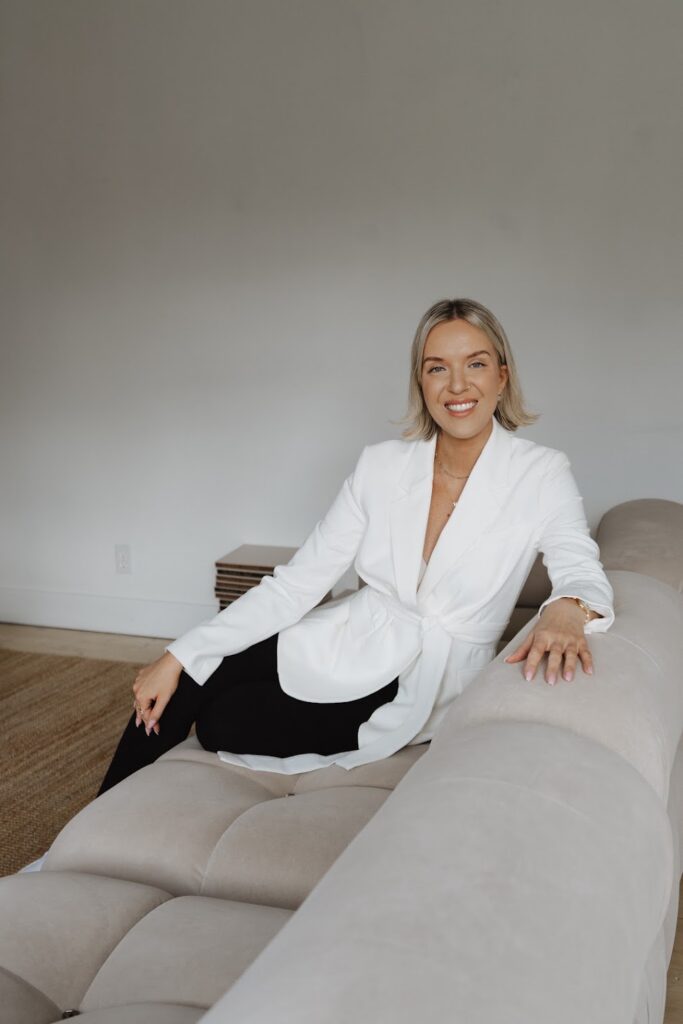 Personal Brand and PR Expert Katrina Owens sits on a nude couch in a white room smiling at a camera looking towards the side of the couch in black pants and a white blazer