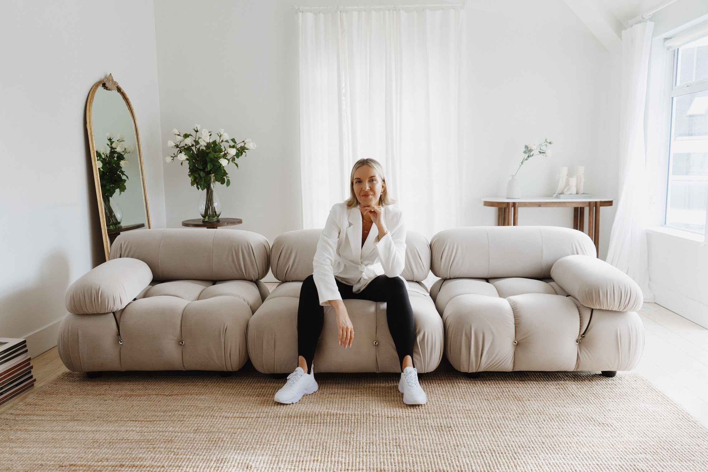 Katrina Owens, who teaches personal brands how to land brand partnerships, sits on a couch in a white room wearing a white blazer, black pants, and white sneakers