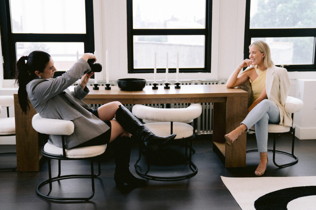 Speaker, author, and podcaster Katrina Owens sits at the end of a table while her photographer sits at the other end taking photos of her posing in jeans, a butter yellow top, and a white blazer