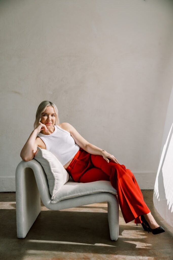 Katrina Owens sits on a neutral chair in a white room wearing a white sleeveless shirt and red pants while teaching about online visibility