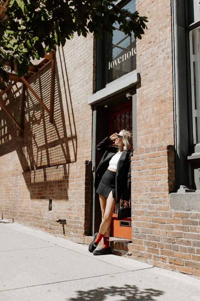 Author Katrina Owens stands in the doorway of a brick building in a black skirt, white shirt, and black jacket wearing sunglasses