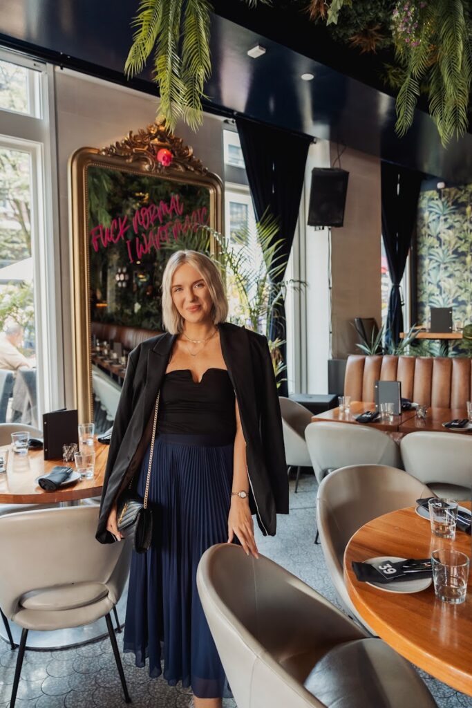 Personal Brand Speaker and Author Katrina Owens stands in a modern trendy restaurant wearing a black blazer, black top and blue skirt 