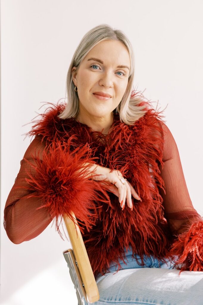 Katrina Owens, Personal Brand Coach for executives, industry leaders, and entrepreneurs, sits in a chair wearing a mesh red top with feather collar and cuffs