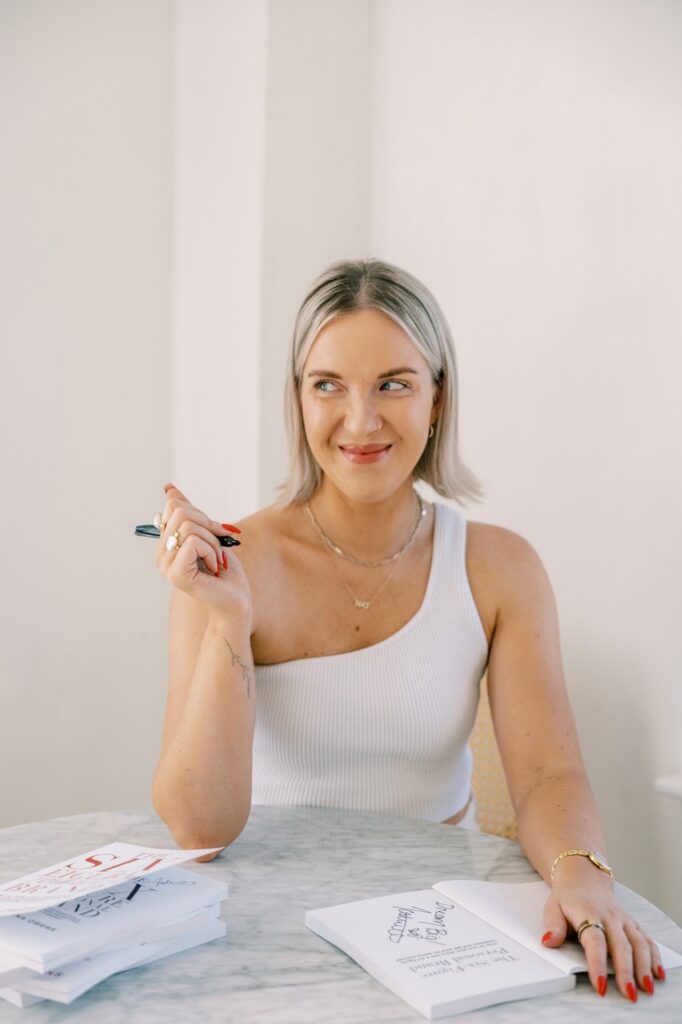 Personal Brand Coach Katrina Owens sits in a chair smiling looking off to the side wearing a one-shoulder white top sitting at a marble table signing copies of her book