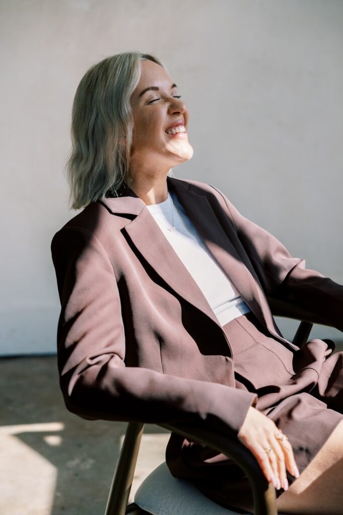 Katrina Owens, who coaches personal branding to executives and entrepreneurs, smiles sitting in a chair looking towards a sunlight window in a white top and black blazer